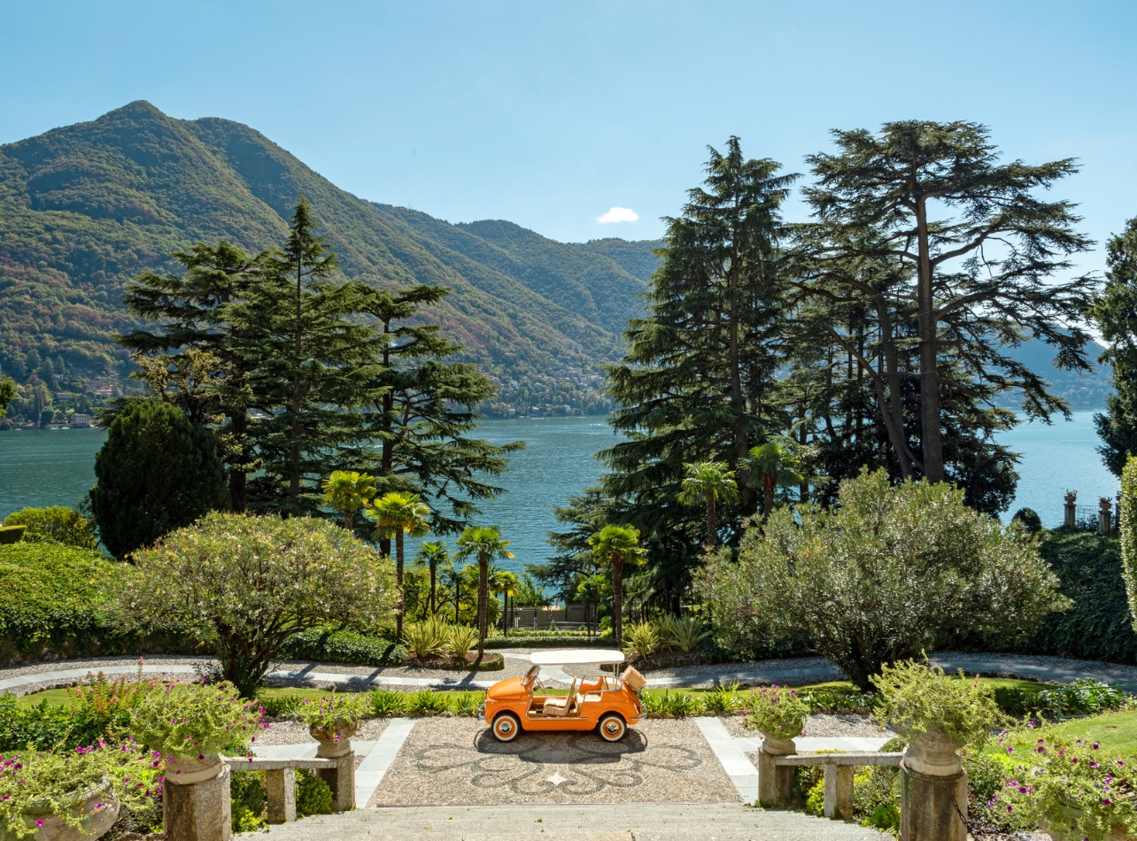 A Fiat 500 Spiaggina in orange parked in front of the Passalacqua hotel surrounded by greenery and water in the background