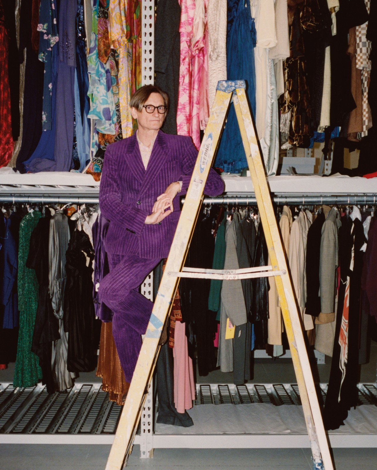 Hamish Bowles standing on a ladder in a purple suit in his fashion archive