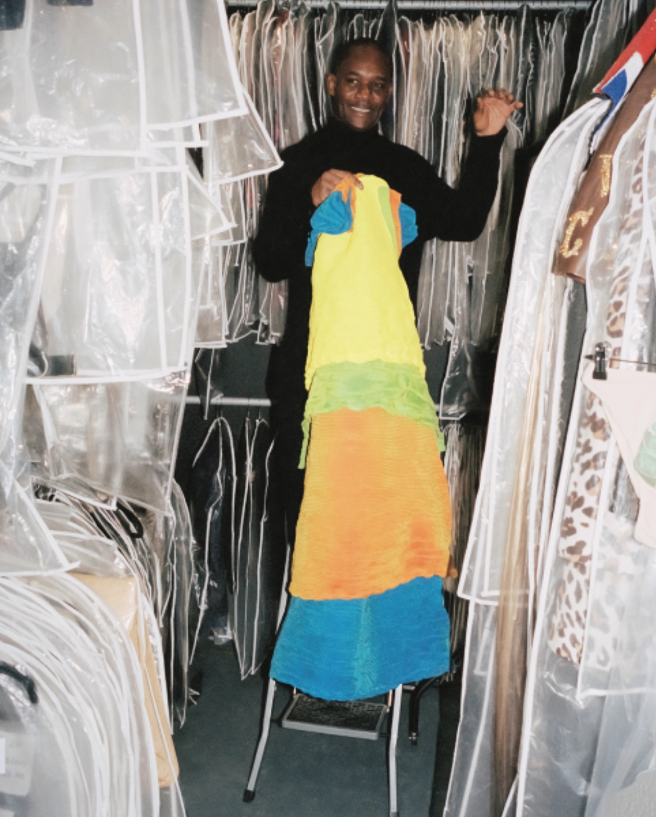 Jefferson Ihenacho standing on a small ladder in the middle of a closet of clothes holding a bright colored dress