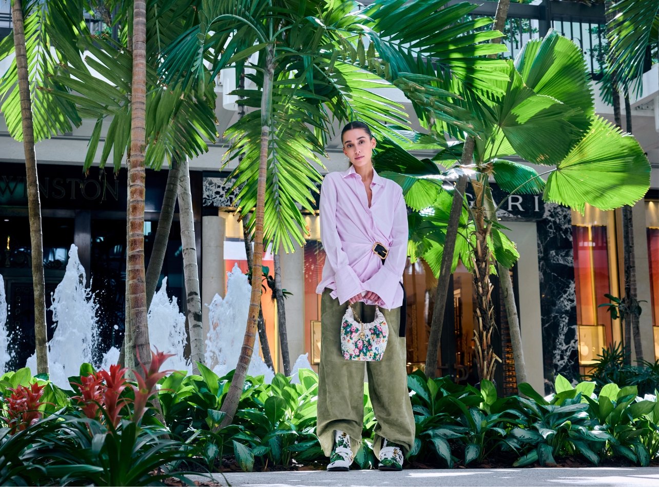 Model wearing Loewe pink belted top and green suede pants holding a beaded bag surrounded by greenery