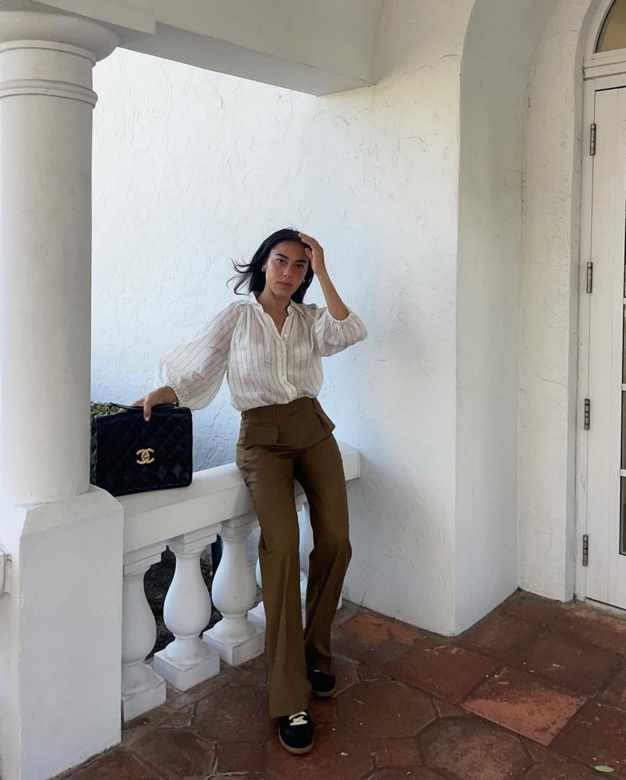 Portrait of Devorah Ezagui wearing brown pants white striped shirt with a large black chanel bag leaning against white railing