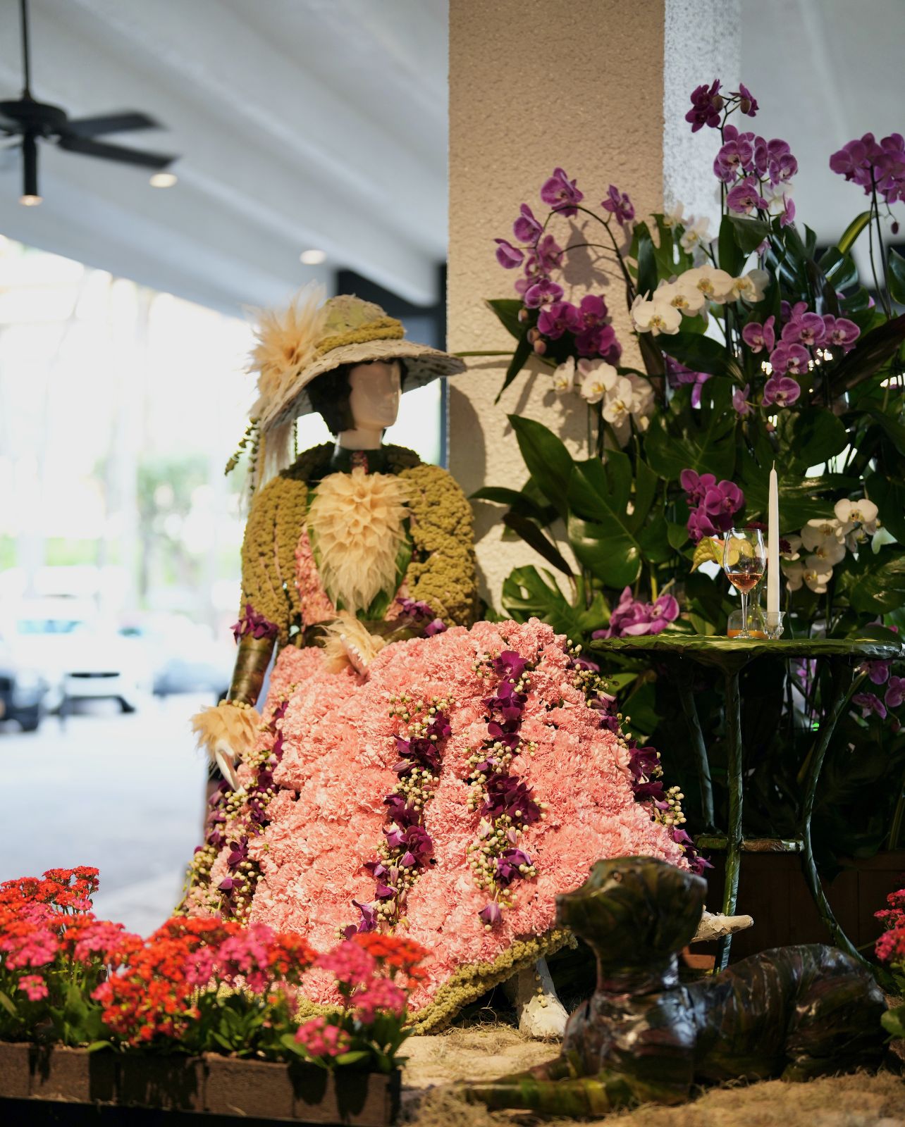 Mannequin of female sitting in a chair with a table with candle and glass of wine. Dress is made out of a variety of pink flowers and a hat made up of moss