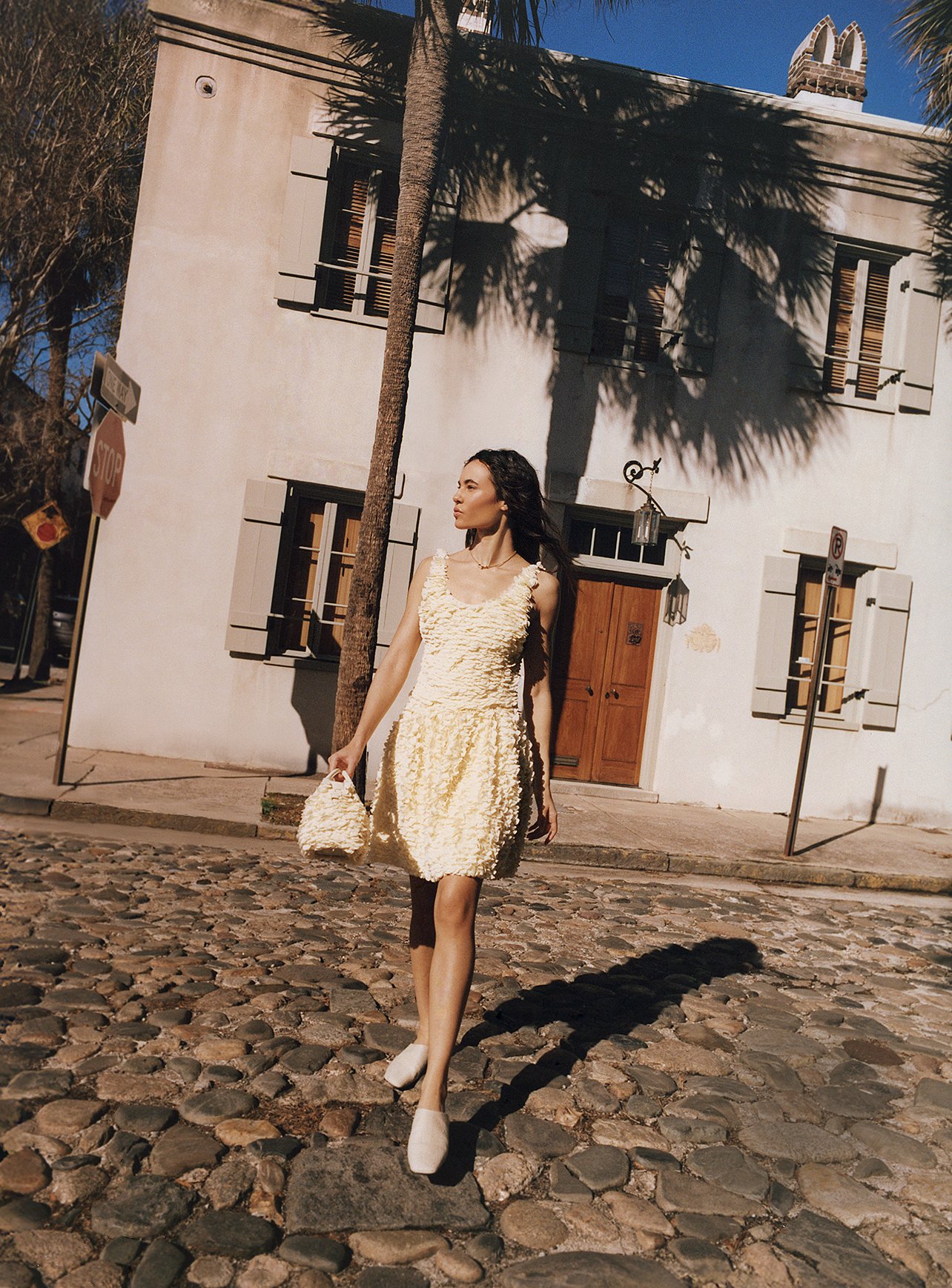 Model wearing a yellow Loro Piana dress with handbag walking across a cobblestone street