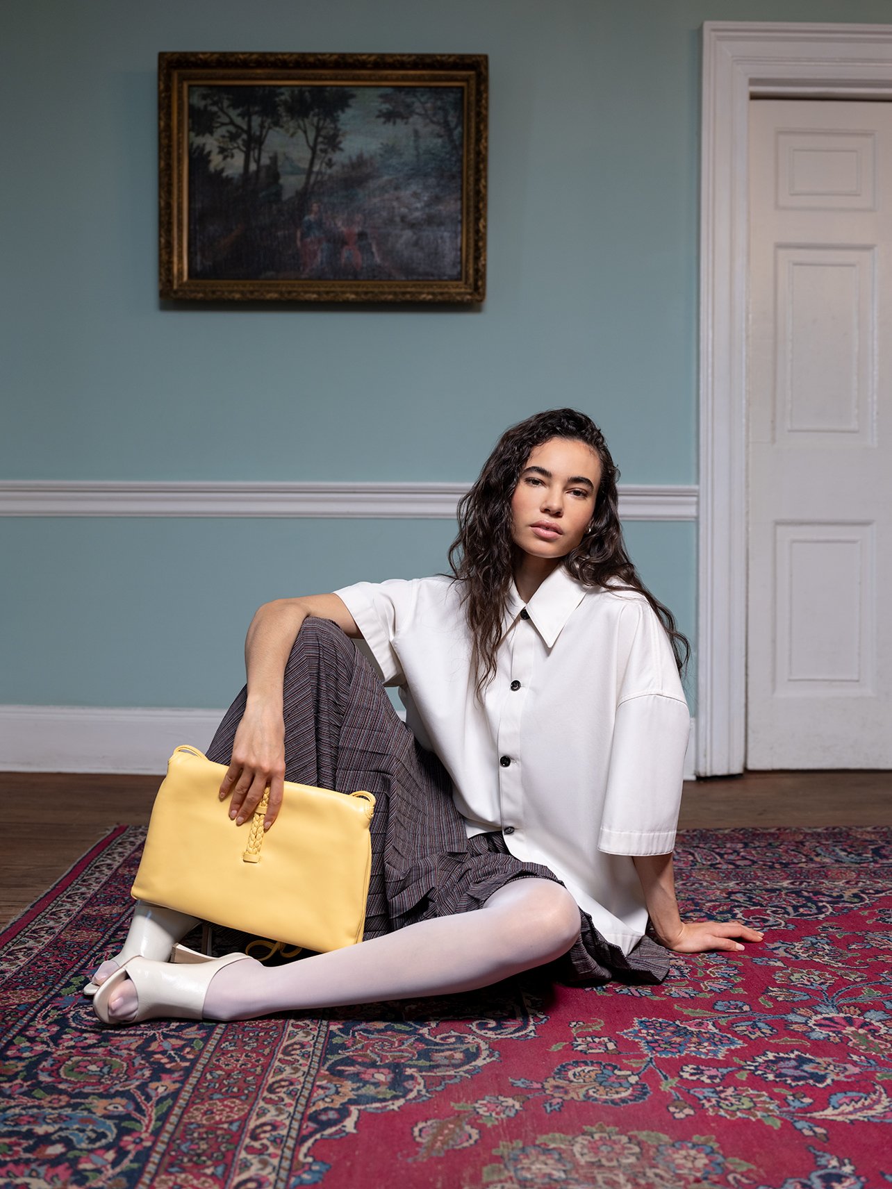 Model wearing a full Bottega Veneta look with white skirt, gray skirt and yellow handbag sitting on a red printed rug and blue walls