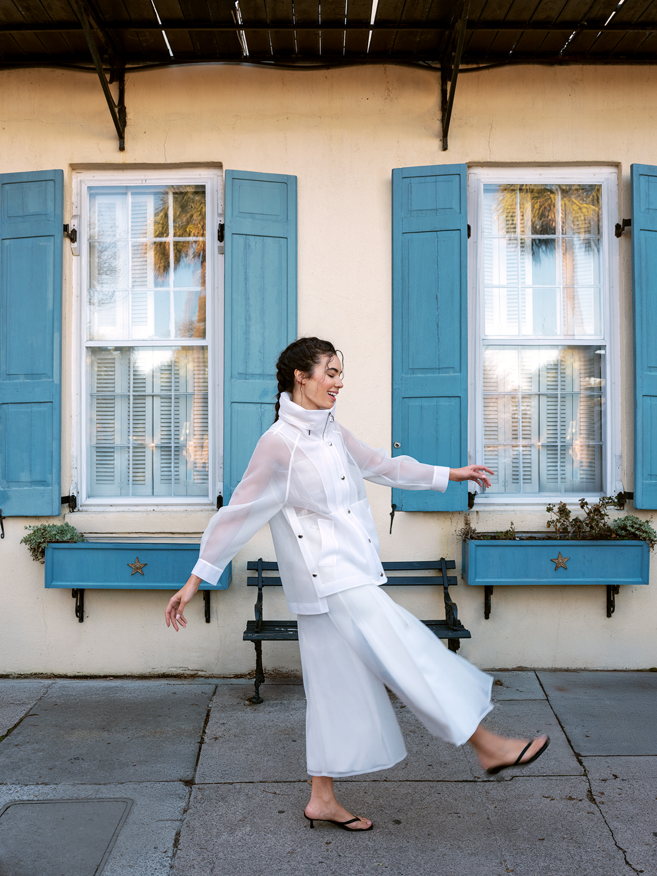 Model wearing a white Akris jacket and pants with Toteme black sandals with blue shuttered home in the back